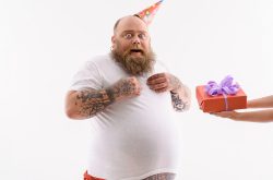 Fat man is receiving gift box. He is standing and looking at camera with surprise. Man is wearing birthday cone. Isolated